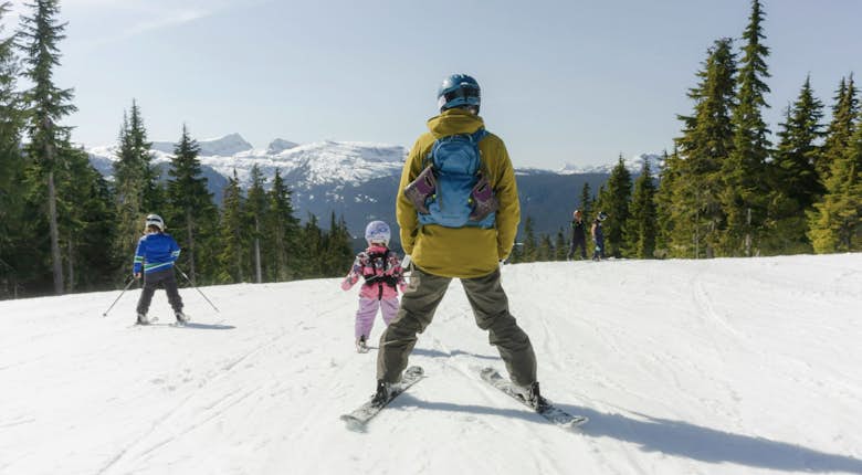A person assists a child skiing down a snowy slope, surrounded by pine trees and distant snow-capped mountains.