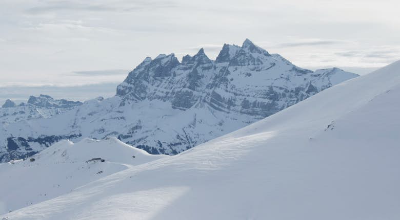 Snow-covered mountain range with sharp peaks in the background and a gently sloping snowy hill in the foreground, under an overcast sky.