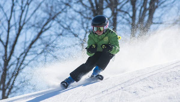 A skier in a green jacket and helmet descends a snowy slope, with trees in the background.
