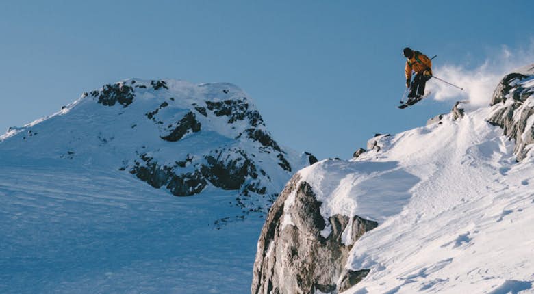 A skier in an orange jacket jumps off a snow-covered cliff with a snowy mountain peak in the background on a clear day.