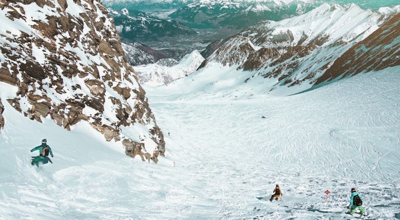 Three people skiing down a snow-covered mountain slope with rocky cliffs, surrounded by snow-covered peaks and a valley in the background.