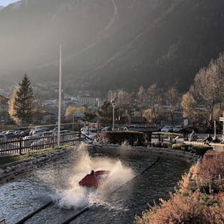 A person on a jet ski performs a trick in a water channel with an autumnal mountain town in the background during sunset.