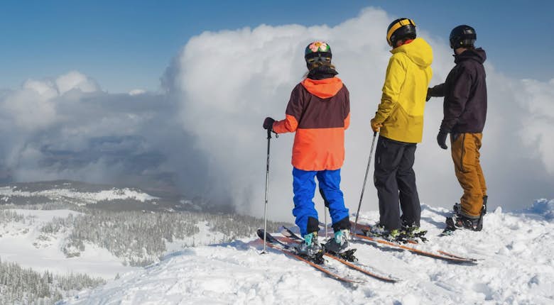 Three skiers in bright gear stand on a snowy mountain peak, looking at a cloudy sky in the distance.