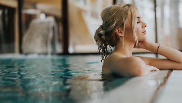 A woman with blonde hair relaxes with her eyes closed in an indoor pool, leaning on the edge.
