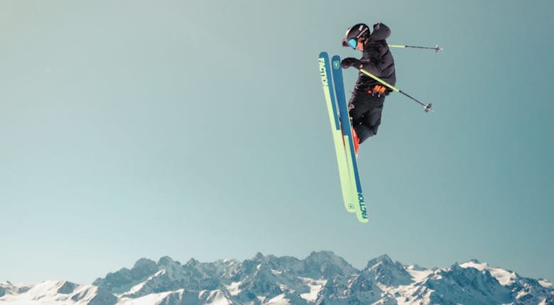 A skier performing an aerial trick in mid-air with alpine mountains in the background under a clear sky.
