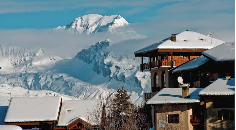 Snow-covered houses with wooden balconies in the foreground and a majestic, snow-capped mountain peak under a clear blue sky in the background.