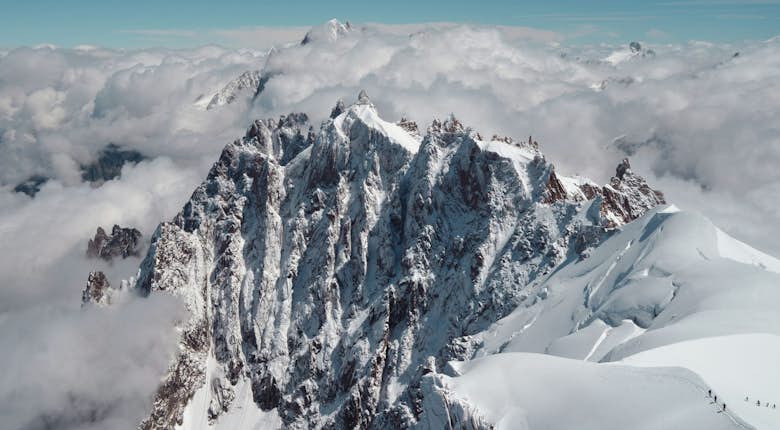 Snow-covered mountain peaks rise above the clouds under a clear blue sky. A group of climbers is visible on the right side of the image.