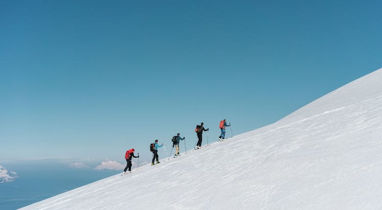 A group of six people equipped with skiing gear ascends a snow-covered slope under a clear blue sky.
