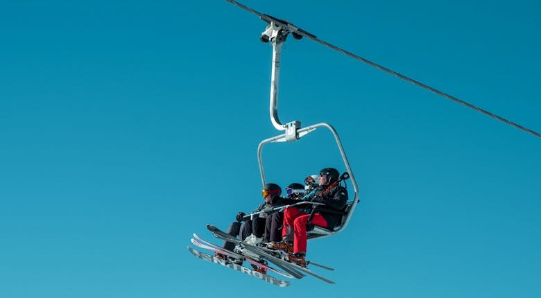 Two skiers on a chairlift against a clear blue sky, with ski equipment attached, heading up a mountain.