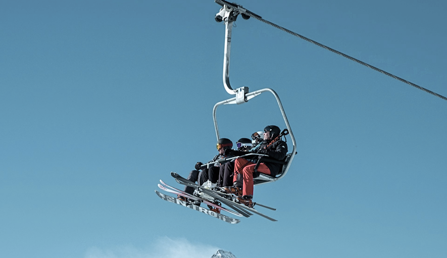 Three people in ski gear on a chairlift with a snowy mountain peak in the background under a clear blue sky.