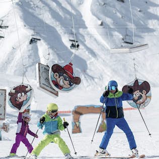 Children in colorful ski gear learn to ski using a themed training course with cartoon characters on a snowy mountain.