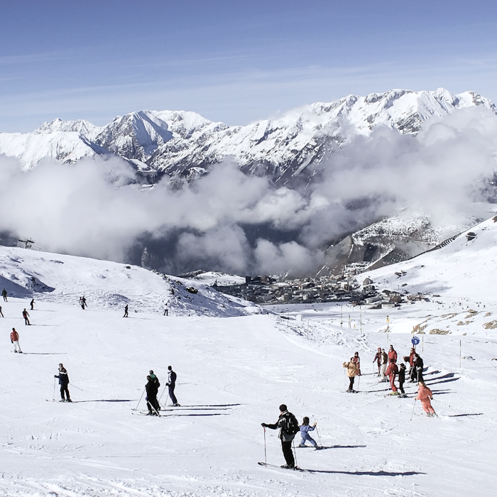 Skiers on snowy slopes with a backdrop of majestic mountains and clouds, with a small village visible below.