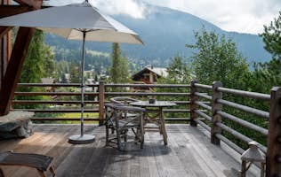 Verbier - 4 Verbier accommodation - Chalet Jenna - A wooden outdoor table with wicker chairs, a vase of white flowers, two green candle holders, and an open book on a balcony overlooking a green mountainside.