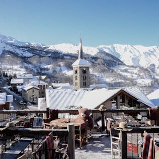 View from a snowy rooftop terrace overlooking a quaint village with a church spire, surrounded by snow-covered mountains under a clear blue sky.