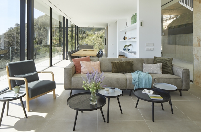 Casa Pere, Costa Brava Modern living room with a beige sofa, colorful cushions, round coffee tables, an armchair, large windows, and a view of greenery outside.
