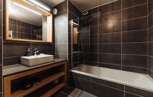 Megeve accommodation - Apartment Opale - Modern bathroom interior with a bathtub, shower, wooden vanity unit, rectangular mirror, and dark tile walls.