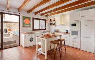 Pollensa - 4 Mallorca accommodation - Ca Na Rieres - Bright dining area with wooden beams, white furniture, and a purple polka-dot rug, adjacent to an open kitchen with tile flooring and access to an outdoor patio.