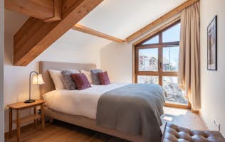 Alpe d'Huez - 2 Baqueira Beret alojamiento - Apartamento Écrin - A cozy bedroom with a double bed, gray and mauve cushions, bedside lamp, angled wooden ceiling, large window with beige curtains, and a view of snowy buildings outside.