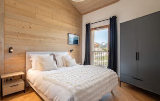 Alpe d'Huez - 2 Alpe d’Huez accommodation - Apartment Nivalis - A modern bedroom with a wooden accent wall, a neatly made bed with white linens, a dark wardrobe, a nightstand, and a window with black curtains overlooking an outdoor view.