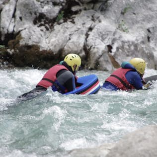 Two people in helmets and life jackets paddling a blue inflatable raft through turbulent white-water rapids, surrounded by rocky banks.