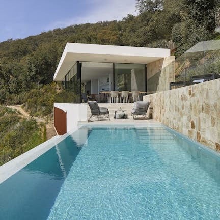 Modern house with large glass doors overlooking an infinity pool and wooded hillsides.