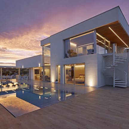 Modern two-story house with large windows and a swimming pool, illuminated at twilight under a colorful sky.