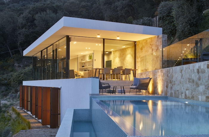 Casa Pere in Costa Brava Modern glass-walled house with illuminated interiors, overlooking an infinity pool and surrounded by trees at dusk.