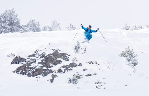 A skier in blue jumps over a rocky patch on a snowy slope, amid a woodland landscape covered in snow.