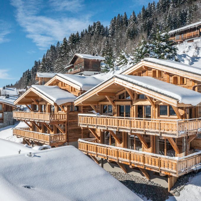 Les Gets property management by Emerald Stay Several wooden chalets covered in snow are situated on a mountainside, surrounded by a snowy landscape and evergreen trees, under a clear blue sky.