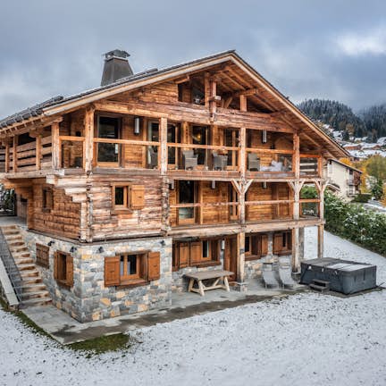 Traditional wooden chalet with stone foundation in a snowy landscape, surrounded by scattered snow and autumnal trees.