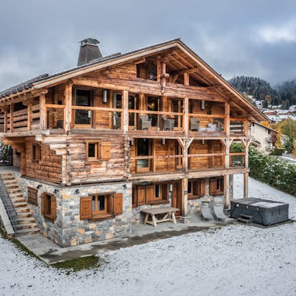 Traditional wooden chalet with stone foundation in a snowy landscape, surrounded by scattered snow and autumnal trees.