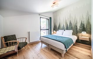 Chamonix alojamiento - Apartamento Edelweiss - Modern bedroom with a double bed, forest mural on the wall, wooden floor, armchair, and large window letting in natural light. Chamonix alojamiento - Apartamento Edelweiss - Modern bedroom with a double bed, forest mural on the wall, wooden floor, armchair, and large window letting in natural light.