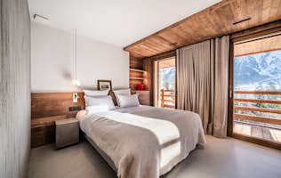 Les Houches - 3 Chamonix location - Chalet Foxden - A cozy bedroom with a wooden interior, featuring a double bed, soft bedding, pendant lamp, side table, and large windows showcasing a snowy mountain view.