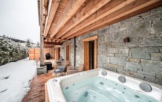 Châtel - 4 Morzine alojamiento - Apartamento Ubuntu - Wooden deck with stone walls, featuring two wicker chairs, a round table with a teapot and cups, and a wooden hot tub topped with folded towels. Roof beams and a fenced backdrop complete the scene.