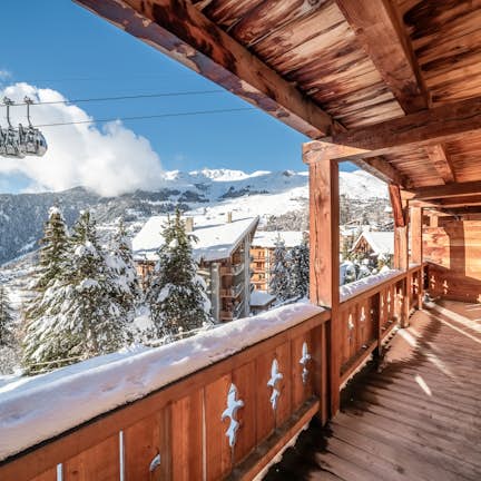 View from a wooden balcony overlooking a snowy mountain landscape with a cable car passing overhead and chalets in the background.