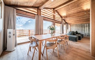 Les Houches - 2 Chamonix location - Chalet Foxden - Wooden dining area with a table and chairs, large windows, and a view of snow-capped mountains. Natural light illuminates the room.