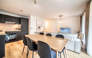 Les Gets - 2 Chamonix alojamiento - Apartamento Dolmen - Modern living and dining area with a wooden table and black chairs. The kitchen has black cabinets. A TV is on, and there's a painting of a deer on the wall. The space is well-lit with natural light.