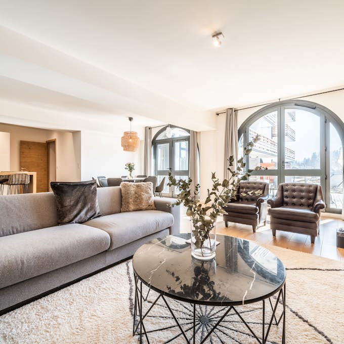 Service de conciergerie à Chamonix  Conciergerie Chamonix Spacious living room with a large gray sectional sofa, a round marble coffee table, and two armchairs by arched windows. The room features wooden accents and an adjoining open kitchen area.