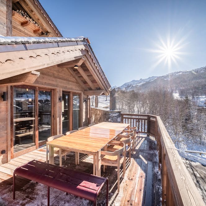 Méribel - 1 Méribel location - Chalet Vallon - Wooden terrace with a long table and chairs, overlooking a snowy mountain landscape under a bright sun.
