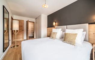 Alpe d'Huez - 3 Verbier alojamiento - Apartamento Koya - Modern bedroom featuring a large bed with white bedding and gold pillows, wooden accents, a mirrored closet, and minimalistic hanging light.