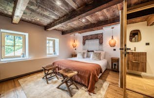 Morzine - 2 Morzine accommodation - Chalet Le Soli - A cozy bedroom with wooden beams, a double bed with a brown throw, two wooden stools at the foot, two windows, pendant lights, and an attached bathroom with a wooden vanity.