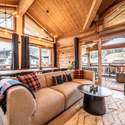 Apartment Pamir, Les Gets Cozy wooden chalet living room with large windows, a beige sofa, plaid pillows, a black coffee table, and a view of neighboring houses and trees.