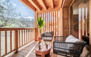 Les Gets - 4 Les Gets location - Appartement Colibri - A sunny balcony with wooden beams, featuring a black wicker chair, a round table with plants, and a view of green trees and blue skies.