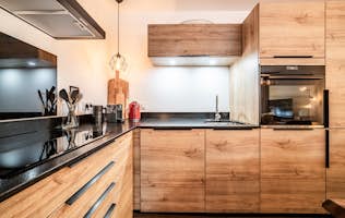 Châtel - 3 Morzine alojamiento - Apartamento Ubuntu - Modern kitchen and dining area with a wooden table, and kitchen appliances. Wooden accents and a framed picture on the wall. Warm lighting fixtures are overhead.