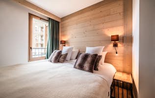 Alpe d'Huez - 3 Alpe d’Huez accommodation - Apartment Tineo - A cozy bedroom features a large bed with white linens, several pillows, and dark cushions. Two wall-mounted lamps provide warm lighting on a wooden accent wall. A window with a curtain is on the left.