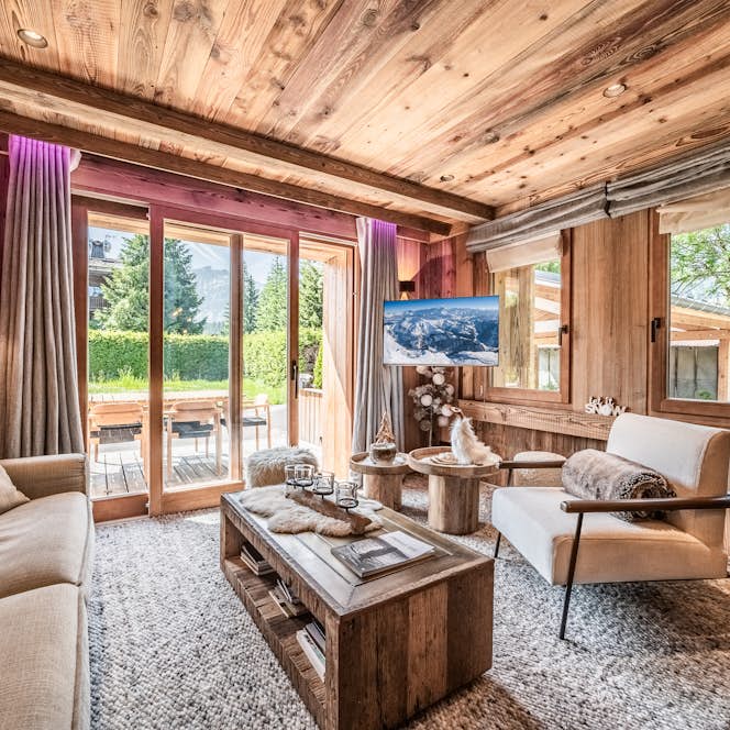 Megève - 1 Megeve accommodation - Apartment Brume - A cozy living room with wood-paneled walls and ceiling, a beige sofa, a wooden coffee table, and large glass doors opening to a sunlit patio with greenery outside.