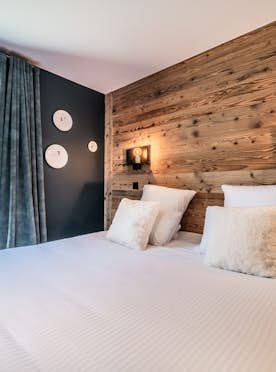 Les Gets location - Appartement Colibri - A cozy modern bedroom featuring a large bed with white linens and fluffy pillows, wooden walls, mounted wall lights, and a window showing outdoor greenery.