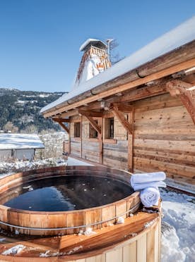 Saint-Gervais - 5 Meribel alojamiento - Chalet Betula - A wooden cabin with a chimney and a hot tub is surrounded by snow, set against a mountainous landscape under a clear blue sky.