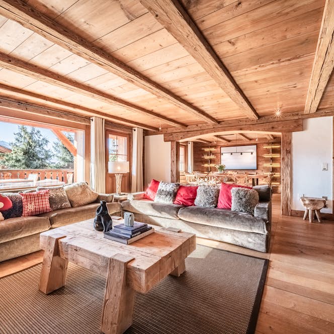 Verbier - 1 Verbier accommodation - Apartment Snowcrest - Spacious wooden living room with two sofas, a fireplace, and large windows offering a view of trees and mountains outside. Cozy decor includes a rug and cushions.