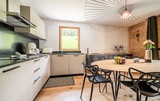 Megève - 4 Megeve accommodation - Apartment Lupin - A modern kitchen with white cabinets, a circular wooden dining table, black chairs, and a large decorative ceiling light. A window allows natural light in and a decorative panther sculpture is on a sideboard.